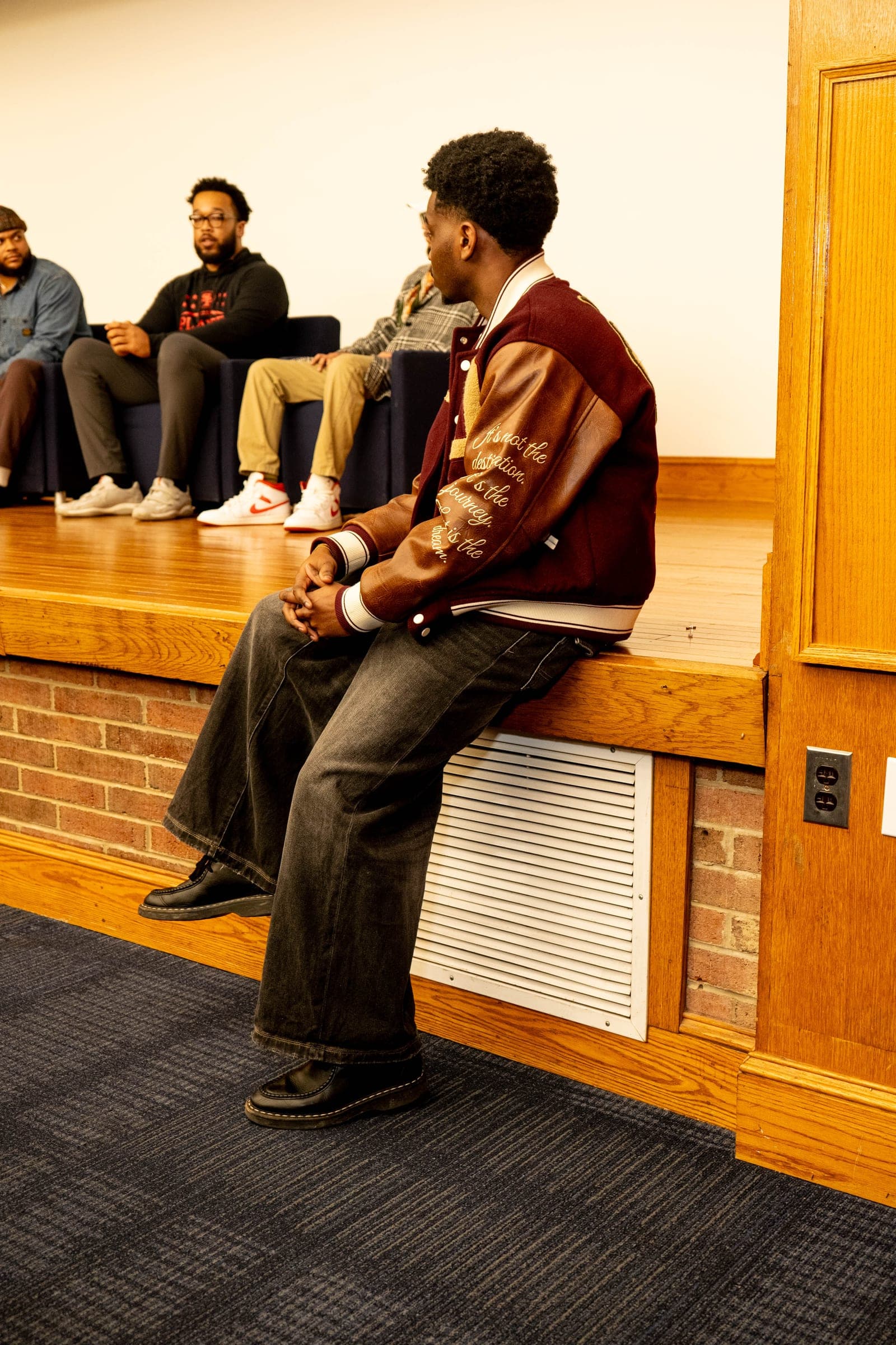 A student in a maroon and brown varsity jacket sits at the edge of the stage, listening as panelists speak behind him.