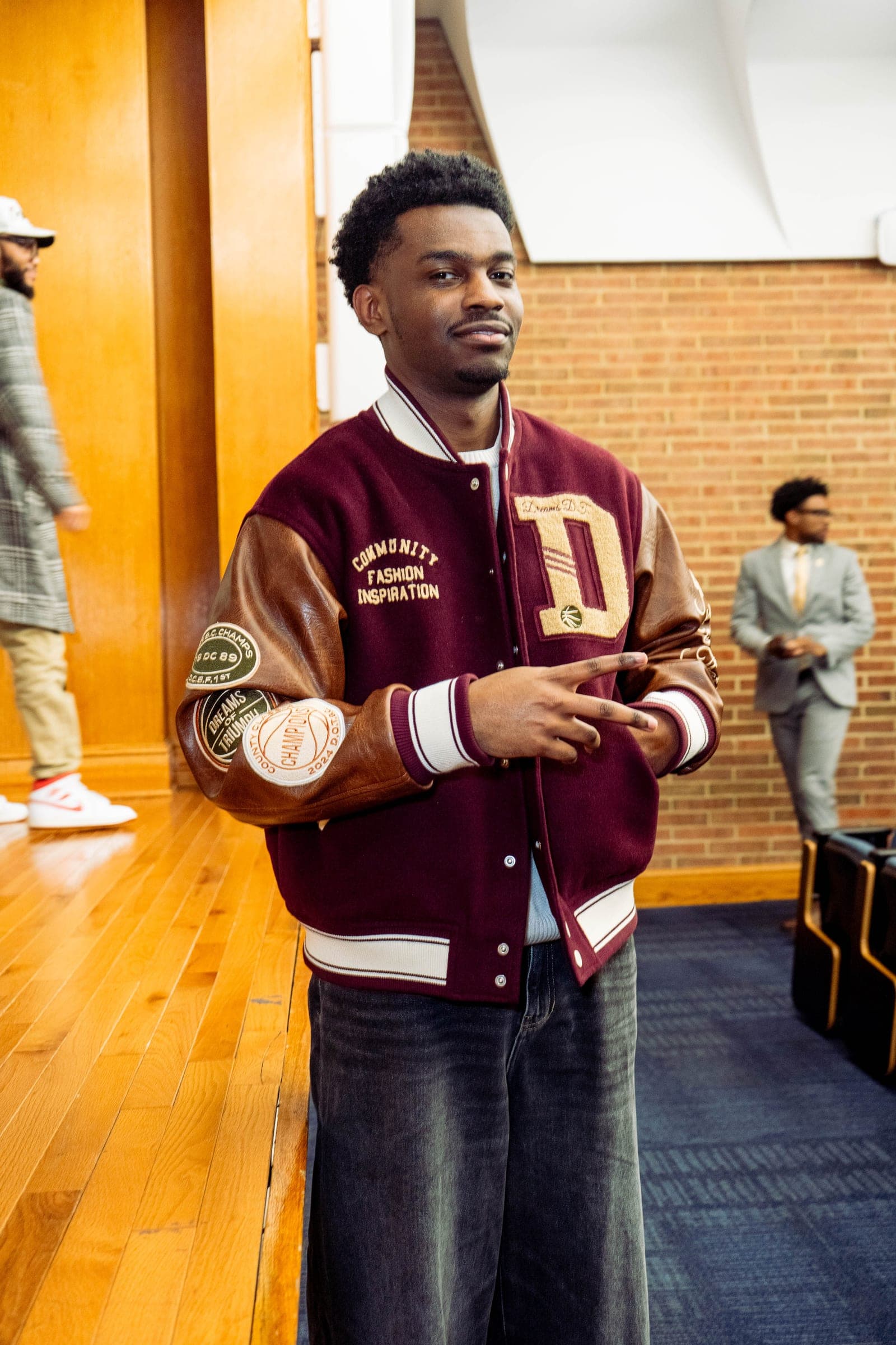 A student in a varsity jacket with 'Community Fashion Inspiration' and 'Dreams of Triumph' patches, smiling toward the camera.